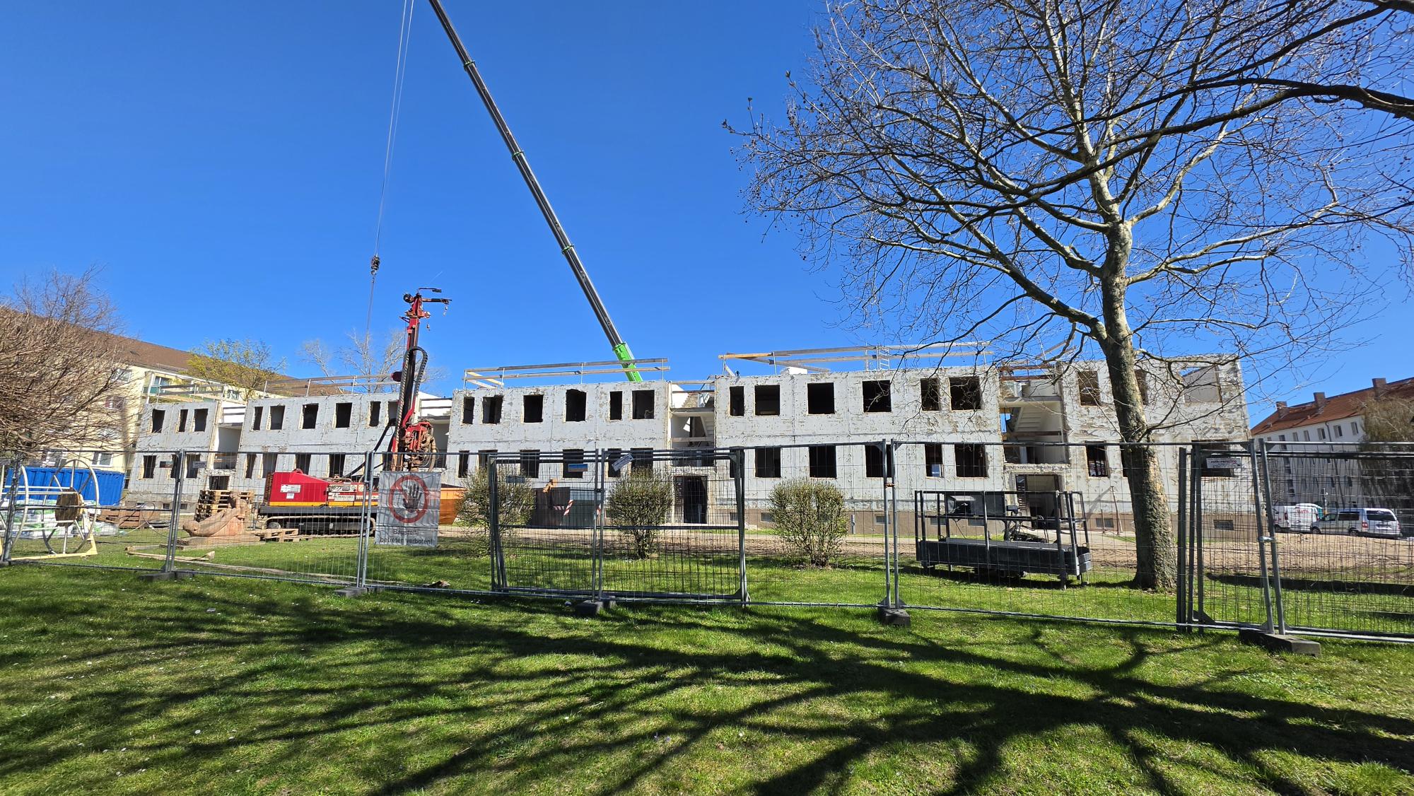 Baustelle vor einem Wohngebäude welches sich im Rohbau befindet, Baumaterialien und Bauzäune sind im Vordergrund.
